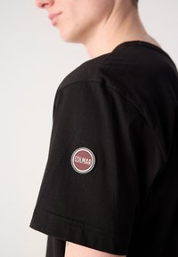 Black cotton t-shirt featuring a round Colmar logo patch in burgundy on the left sleeve; short sleeves and a crew neck design.