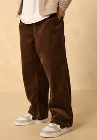 Braune Cord-Hose mit weitem Bein und vertikaler Rippung, getragen mit weißen und grauen Sneakers. Verfügt über seitliche Taschen und einen lockeren Sitz.