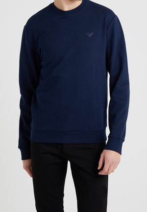 Mann trägt ein marineblaues Sweatshirt mit langen Ärmeln und kleinem dunkelgrauem Logo auf der Brust, kombiniert mit schwarzer Hose.