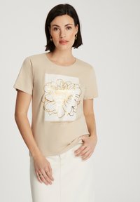 Beżowa koszulka z krótkim rękawem z białym prostokątnym nadrukiem kwiatowym obramowanym złotem. Miękka tkanina o luźnym kroju. Minimalistyczny design.