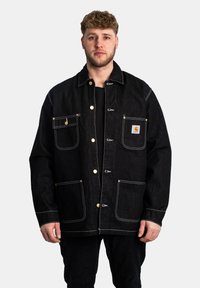 Chaqueta de mezclilla negra con acentos amarillos, grandes bolsillos frontales y costuras blancas en contraste. Cuenta con botones a presión y un diseño de corte relajado.