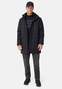Mørkeblå parka med lynlås foran, to store påsyede lommer og en brystlomme. Båret over en mønstret sweater, parret med mørke jeans og sorte sko.