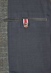 Texturiert grauer Stoff mit einem navyblauen Abschnitt, der eine obere Tasche mit schwarzer Kante und einem Knopf aufweist, akzentuiert durch ein rot-weißes Band.