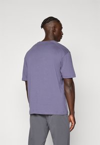 Fioletowy bawełniany t-shirt z krótkim rękawem i okrągłym dekoltem, charakteryzujący się luźnym krojem i minimalnymi detalami szwów, noszony z szarymi spodniami.