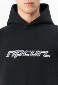 Gros plan d'une personne portant un sweat à capuche noir avec le logo blanc "ripcurl" brodé sur la poitrine.