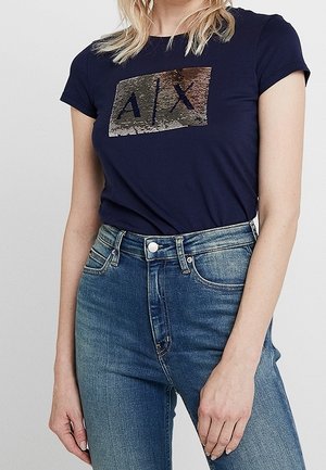 Kvinde iført en mørkeblå t-shirt med et palietbesat A|X-logo, kombineret med højtaljede blå jeans, der er beskåret ved midten af overkroppen og hofterne.