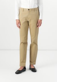 Hordható, egyenes szabású, pamut keverékből készült khaki chino nadrág. Gombos elülső részével, övhurkákkal és két oldalsó zsebével rendelkezik. Fekete balerina cipővel viselik.