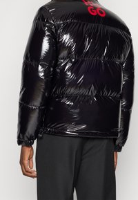 Błyszcząca czarna kurtka puchowa z elastycznymi mankietami i czerwonym napisem na górnej części pleców, noszona na czarnych spodniach, pokazana od tyłu na osobie.