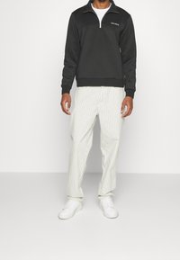 Hombre vistiendo un sudadero negro con media cremallera, pantalones blancos de rayas y zapatillas blancas, de pie contra un fondo blanco liso.