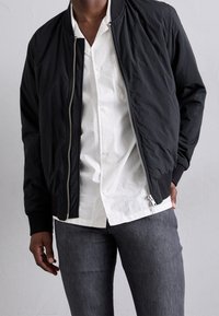Svart bomberjacka med en mjuk tygkvalitet, ribbade ärmslut och nederkant, silverdragkedja, bärs ovanpå en vit skjorta med knappar och grå jeans.