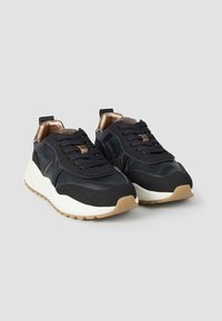 Schwarze Sneakers mit einer glatten, glänzenden Oberfläche, strukturierten Stoffdetails, dicker weißer Sohle und gemusterten schwarzen Schnürsenkeln. Bronzeakzente im Innenbereich.