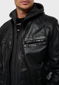 Chaqueta de cuero negra con dos bolsillos en el pecho y cierre de cremallera, superpuesta a un sudadero con capucha gris. Presenta un acabado texturizado y costuras resistentes.