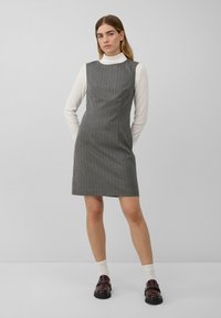Jauna sieviete, tērpta pelēkā slēgtā kleitā ar svītrām, virs baltā turtleneck, ar baltām zeķēm un tumši brūnām mokasīnām, stāv uz vienkrāsaina fona.