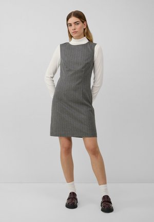 Junge Frau in einem grauen, gestreiften ärmellosen Kleid über einem weißen Rollkragenpullover, weißen Socken und dunkelbraunen Slippern, die vor einem einfarbigen Hintergrund steht.
