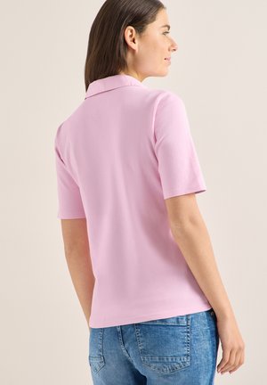 Femme portant un polo rose clair à manches courtes et un jean bleu, vue de dos devant un fond uni.