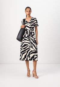 Svart-hvitt stripete kjole med korte ermer, V-hals og knyttet midje. Modellen holder en svart tote bag og har på seg nude blokkhevede sandaler.