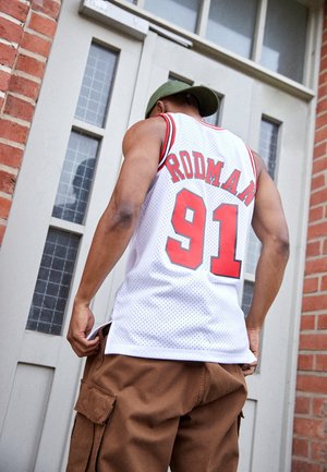 Hombre con camiseta blanca de baloncesto Rodman número 91 y gorra verde, de pie junto a una puerta gris cerrada con paredes de ladrillo.
