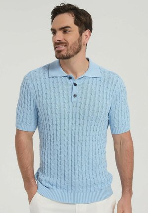 Homme aux cheveux courts et barbe portant un polo en maille texturée bleu clair et un pantalon blanc, debout les mains dans les poches, regardant vers la gauche.