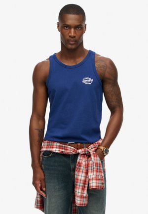 Mørkeblå bomulds tanktop med rund halsudskæring og logo på brystet, parret med prikket flannel bundet om livet og denim jeans.