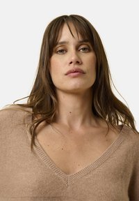 Beige gestrickter Pullover mit V-Ausschnitt, der eine weiche Textur und eine lockere Passform aufweist. Der Stoff wirkt leicht und gemütlich.