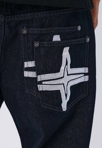 Jeans in denim scuro con un grande disegno bianco ricamato sulla tasca posteriore, con due strisce bianche sopra. Elementi metallici visibili.