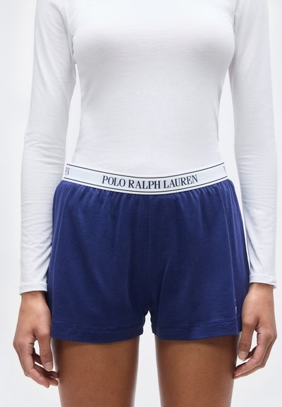 Γυναίκα που φοράει σορτς σε ναυτικό μπλε με άσπρη ελαστική μέση που φέρει τη μάρκα «Polo Ralph Lauren» και λευκό μακρυμάνικο εφαρμοστό τοπ, όρθια.