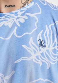 Light blue t-shirt featuring white floral patterns and a contrasting embroidered logo in dark blue. Soft fabric with a crew neckline.