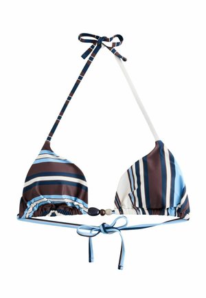 Kobieta z długimi blond włosami, ubrana w paskowany bikini, stoi w pobliżu kamiennego muru, w tle morze i czyste niebo.