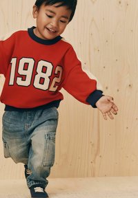 Sweatshirt rouge avec des manches blanches accentuées et le texte "1982". Pantalons cargo en denim bleu avec des poches. Baskets bleues. Fond en bois.