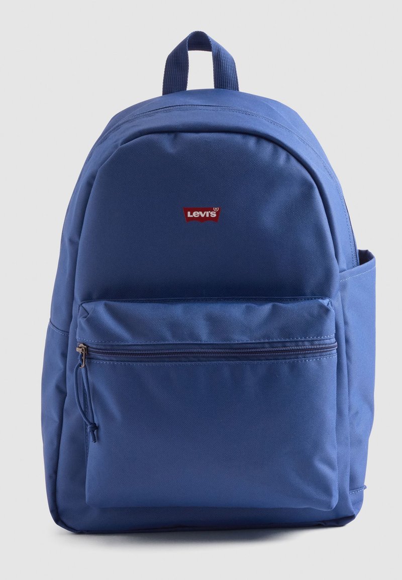 Mochila de tela azul con una textura suave, que presenta un bolsillo frontal con cremallera, etiqueta de logo y un asa superior para llevar.