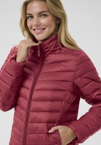 Giacca imbottita rossa con chiusura a zip, cuciture verticali, colletto alto e polsini elastici. Tessuto leggero con texture liscia e lucida.