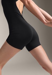 Černý bodysuit s krátkými nohavicemi, který má výřez na zádech a hladký, elastický materiál. Zvýrazňuje tvar těla a pohyb.