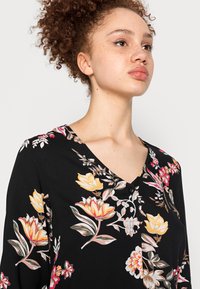 Blusa preta com padrão floral, apresentando flores amarelas e cor-de-rosa, com decote em V e três botões na parte da frente, feita de um tecido leve e texturizado.