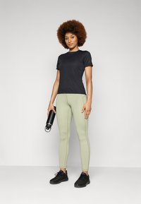 Camisa preta de manga curta, leggings verdes e sapatilhas de desporto pretas. Garrafa de água segurada na mão direita. Tecido suave e design ajustado.