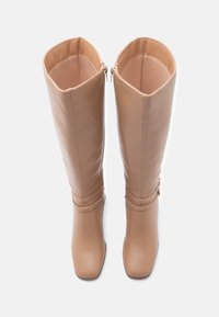 Botas altas de imitación cuero beige con una textura suave, puntera cuadrada, cremalleras exteriores y correas decorativas cerca del tobillo.