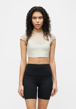 Femeie cu păr creț, purtând un crop top transparent cu mâneci scurte peste un sutien sport alb și pantaloni scurți negri de ciclism cu talie înaltă.