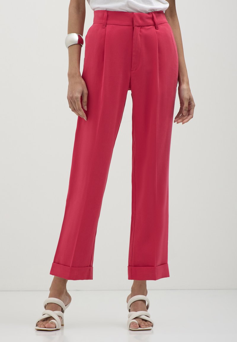 Donna che indossa pantaloni rosa a vita alta e gamba dritta con orli a risvolto, abbinati a sandali con tacco bianchi a fascia e a un braccialetto argentato.