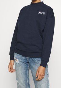 Mörkblå sweatshirt med ribbad halsringning och cuffs, med en liten G-Star Raw Denim-logotyp på bröstet. Bärs med slitna ljusa jeans.