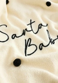 Manta de crema con texto bordado en negro "Santa Baby" y acentos redondos texturizados en negro sobre un fondo de tela suave y esponjosa.