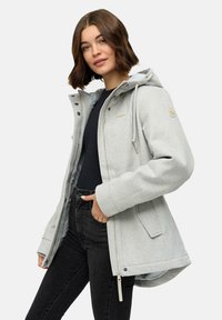 Femme portant une veste à capuche gris clair sur un t-shirt noir et un jean noir, se tenant avec une main dans la poche contre un fond uni.