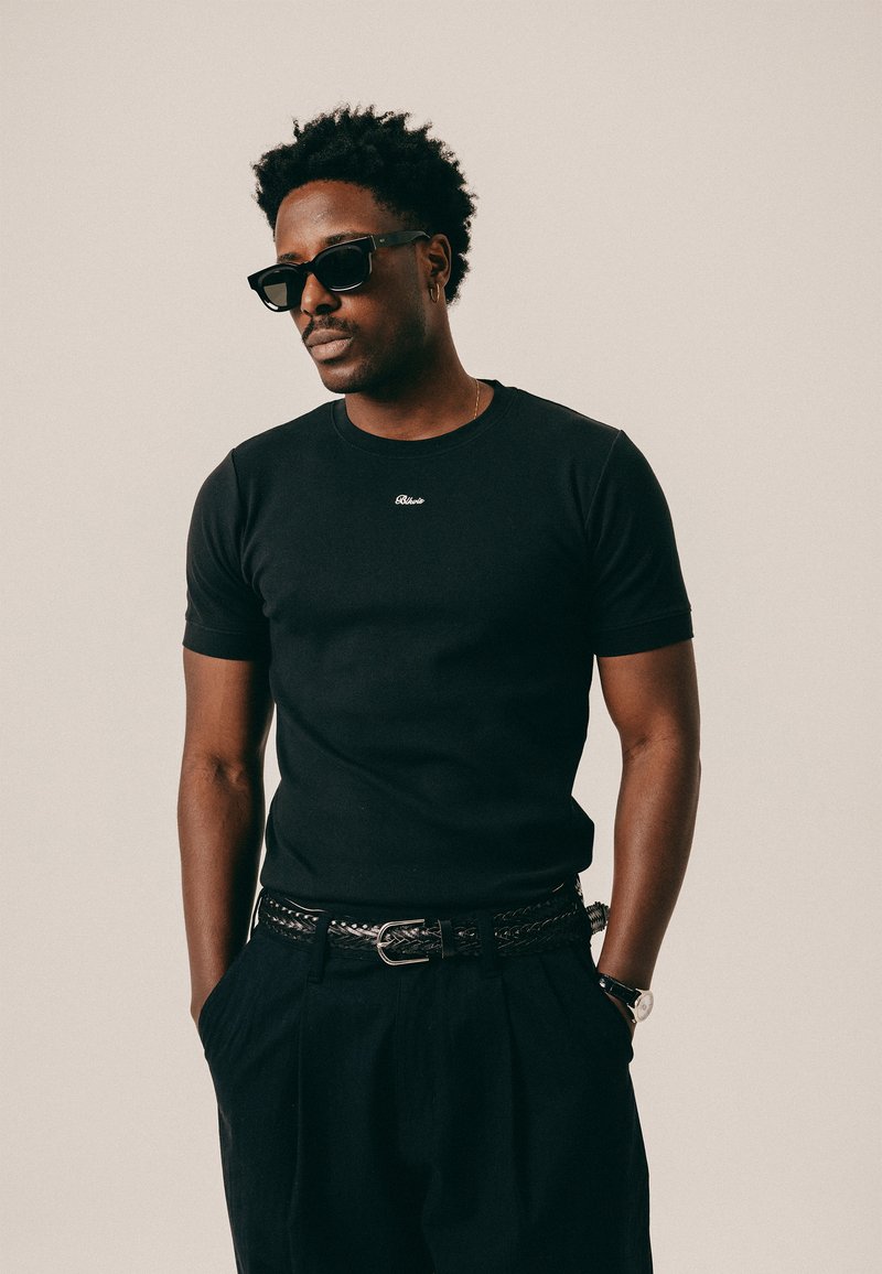 Άνδρας με κοντά σγουρά μαλλιά που φοράει μαύρα γυαλιά ηλίου, εφαρμοστό μαύρο μπλουζάκι, πλεγμένη ζώνη, μαύρο παντελόνι και ρολόι χειρός, στέκεται με τα χέρια στις τσέπες.