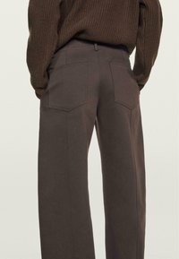 Brune højtaljede bukser med lige ben-design, der har to baglommer og subtilt synlige stikninger. Fremstillet af strukturstof.