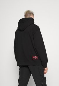 Sweat à capuche noir avec une grande capuche, des poignets et un ourlet côtelés. Comprend un patch rouge dans le bas du dos. Le matériau semble être un tissu doux et texturé.