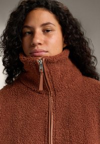 Brown fleece jacket with a high collar and zip closure. The fabric is textured and plush, with a smooth zip pull accent.