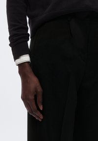 Donkere trui met opgerolde mouwen over een zwarte broek. Close-up van een hand die op de stof rust, waarbij de gladde textuur en vorm worden benadrukt.