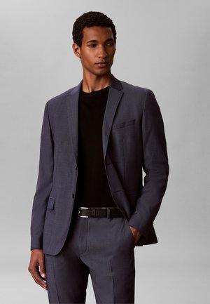 Joven con un traje gris oscuro y una camisa negra, de pie con una mano en el bolsillo frente a un fondo liso gris claro.
