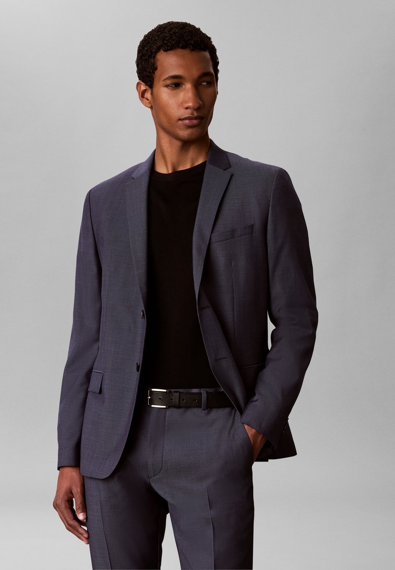 Jeune homme portant un costume gris foncé avec une chemise noire, debout avec une main dans la poche sur un fond uni gris clair.
