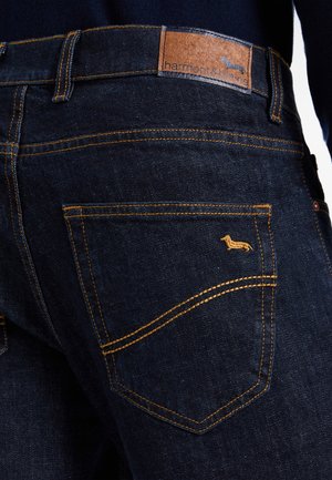 Nahaufnahme einer dunkelblauen Jeanshosen-Hosentasche mit senfgelber Stickerei und einem kleinen Dackel-Logo, das über der Tasche gestickt ist.