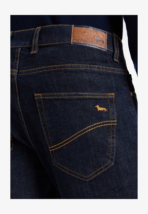 Nærbillede af en baglomme på mørkeblå denimjeans med sennepsgul syning og et lille broderet dachshund-logo ovenover lommen.