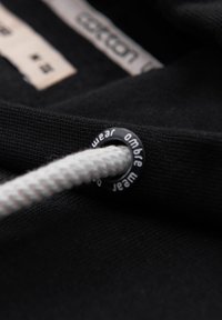 Gros plan sur un tissu noir avec un cordon blanc passé dans un œillet en métal noir portant l'inscription "ombre wear" et des étiquettes de vêtements floues en arrière-plan.